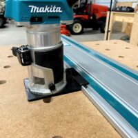 Makita trim router mounted on black adapter base, positioned next to blue guide rail on pegboard workbench