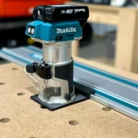 Black guide rail adapter base mounted under a Makita cordless trim router beside a track on an MDF benchtop