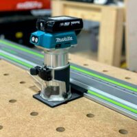 Makita trim router mounted on a clear plastic base adapter, positioned next to a bright green guide rail on a pegboard workbench