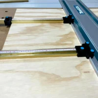 Four black parallel guide brackets with blue knobs mounted on brass T-tracks attached to a Makita track saw guide rail over a wood workpiece