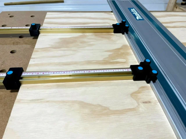 Four black parallel guide brackets with blue knobs mounted on brass T-tracks attached to a Makita track saw guide rail over a wood workpiece