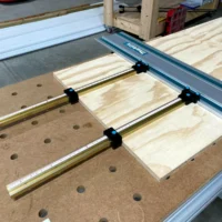 Black ToolCurve parallel guide brackets mounted on brass T-tracks attached to a Makita track saw guide rail, positioned over a pegboard and wood workpieces