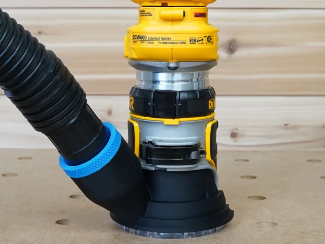 Black hose adapter with blue grip ring attached to DeWalt DCW600 compact router, with black corrugated hose connected to adapter inlet