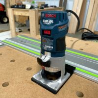 Bosch Colt 1HP router mounted on aluminum guide rail with black plastic adapter base, positioned on cork workbench with green-stripe rail