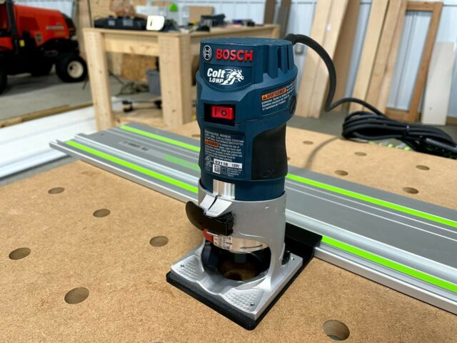 Bosch Colt 1HP router mounted on aluminum guide rail with black plastic adapter base, positioned on cork workbench with green-stripe rail