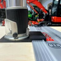 Makita router adapter mounted on a Bosch guide rail, showing the black plastic base attached to the rail with the router positioned above it