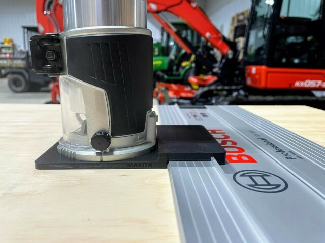 Makita router adapter mounted on a Bosch guide rail, showing the black plastic base attached to the rail with the router positioned above it