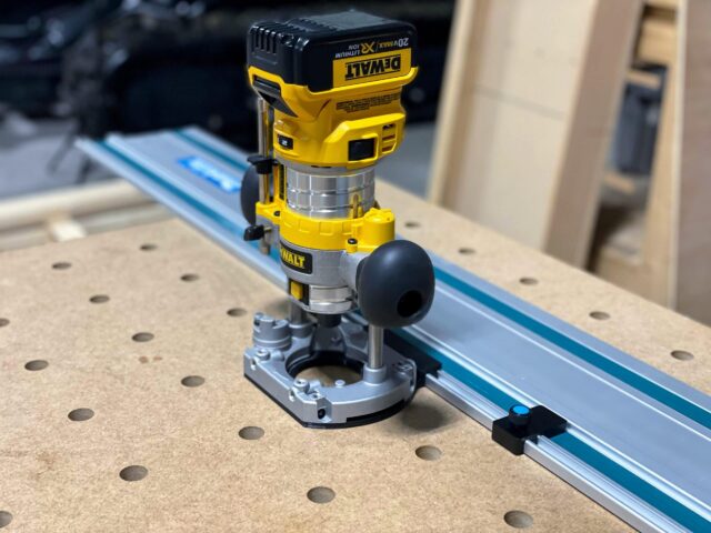 DeWalt trim router mounted on blue Makita guide rail using black and gray plunge base adapter on workbench with pegboard