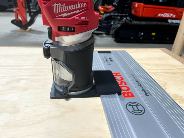 Milwaukee M18 trim router mounted on black adapter plate attached to Bosch guide rail on wooden workbench