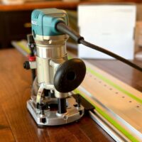 Makita trim plunge router mounted on a ToolCurve guide rail adapter, positioned over a wooden board with a green alignment rail