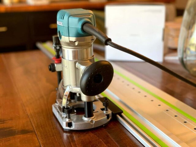 Makita trim plunge router mounted on a ToolCurve guide rail adapter, positioned over a wooden board with a green alignment rail