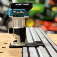 Makita trim router mounted on black adapter plate positioned on Dewalt track saw rail, viewed from the side on a pegboard workbench