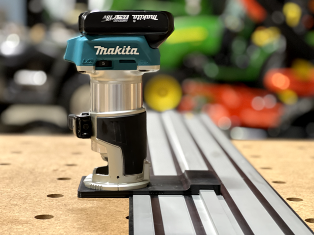 Makita trim router mounted on black adapter plate positioned on Dewalt track saw rail, viewed from the side on a pegboard workbench