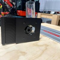 Black plastic Makita router adapter mounted on a Bosch guide rail, showing the threaded center port and four mounting screw holes on the front face