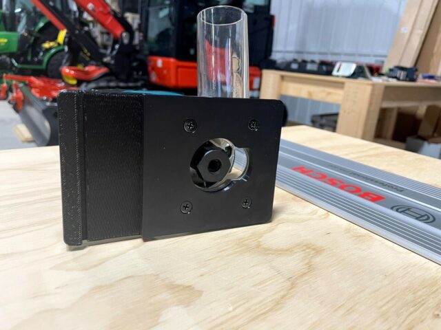 Black plastic Makita router adapter mounted on a Bosch guide rail, showing the threaded center port and four mounting screw holes on the front face