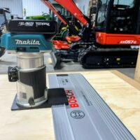 Makita trim router mounted on black adapter plate, positioned on a Bosch guide rail atop a wooden workbench
