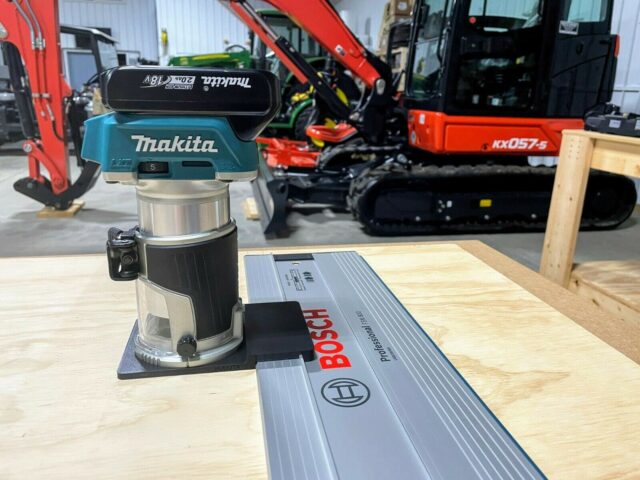Makita trim router mounted on black adapter plate, positioned on a Bosch guide rail atop a wooden workbench