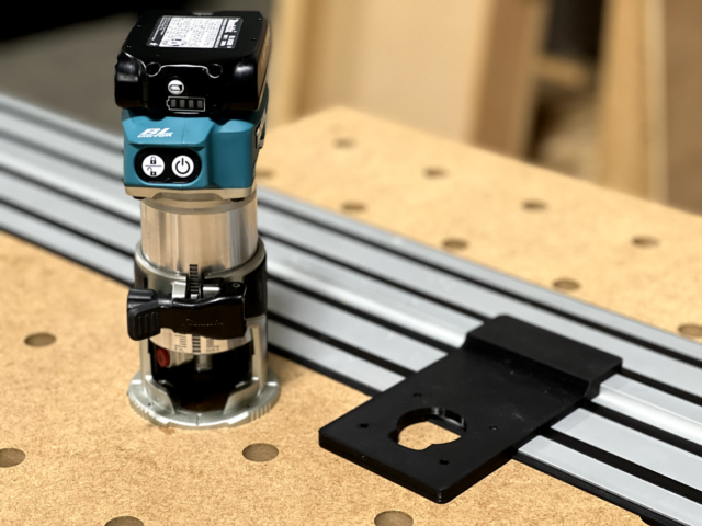Makita trim router mounted on black adapter plate positioned on Dewalt track saw aluminum track over perforated wooden base