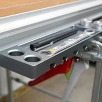 Dark gray utility rack mounted on an aluminum t-track, showing two circular holes for bench dogs and slots holding clamps and tools