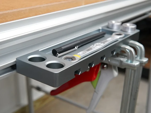 Dark gray utility rack mounted on an aluminum t-track, showing two circular holes for bench dogs and slots holding clamps and tools