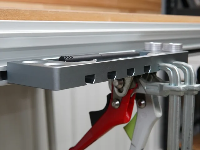 Gray utility rack mounted on aluminum t-track, holding red quick clamps, metal bench dogs, and a black tool in the tray