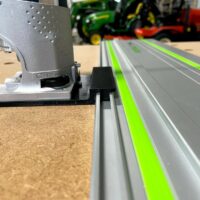 Bosch Colt 1HP router adapter mounted on a Festool guide rail, showing the silver adapter body with perforated face and black mounting bracket engaged with the rail