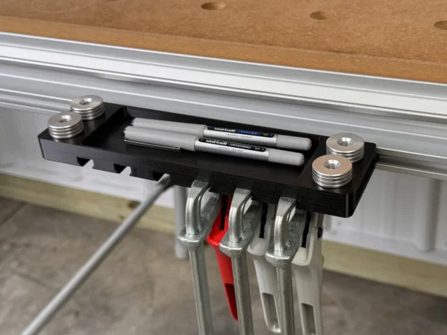 Black utility rack mounted on Festool MFT table track, holding markers and pencils in top tray, with four quick clamps and bench dogs in side slots