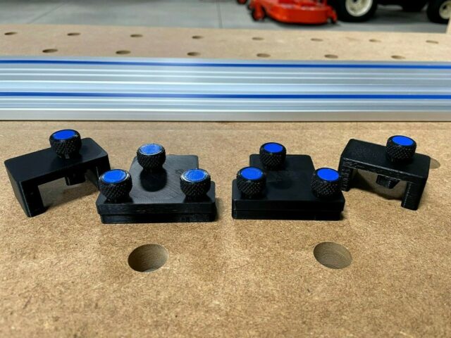 Four black 3D-printed adapters with blue knurled knobs on a perforated workbench surface, positioned above a Kreg guide rail