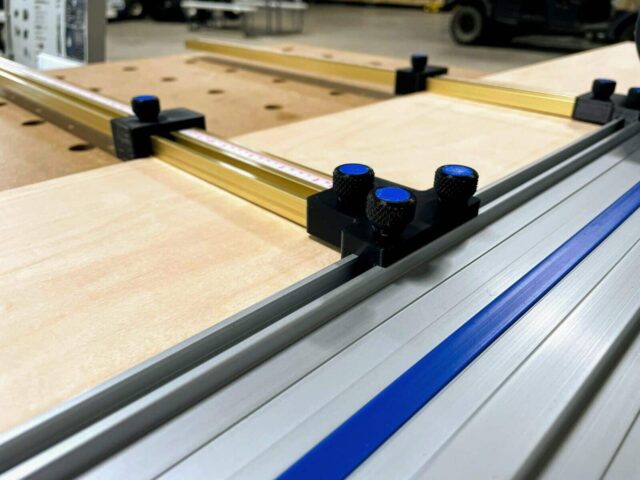 Black plastic parallel guide bracket with three blue knurled knobs mounted on aluminum Kreg ACS guide rail, positioned over plywood workpiece
