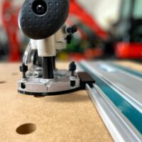 Makita trim plunge router mounted on aluminum guide rail, showing metal baseplate with adjustment screws and black plunge mechanism above