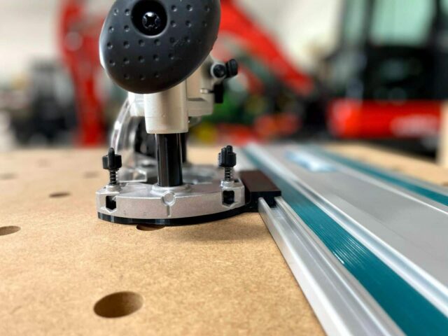 Makita trim plunge router mounted on aluminum guide rail, showing metal baseplate with adjustment screws and black plunge mechanism above