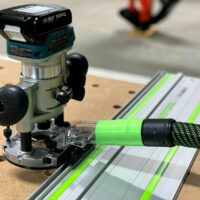 Makita trim plunge router mounted on Festool guide rail with dust hose attached, shown on wooden workbench