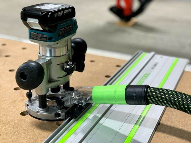 Makita trim plunge router mounted on Festool guide rail with dust hose attached, shown on wooden workbench