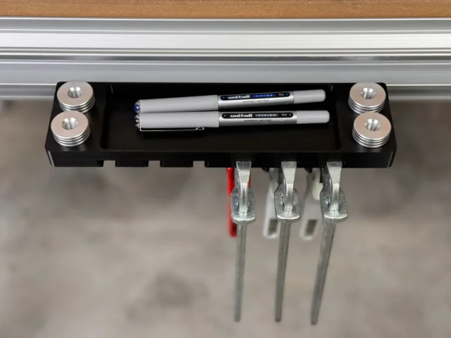 Black utility rack mounted to aluminum t-track, holding two pens and three bench dogs in metal slots, with threaded posts at each corner