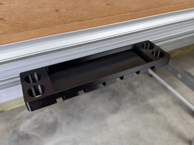 Black plastic utility rack mounted on aluminum t-track, with slots for clamps along the bottom and cylindrical holes on each end