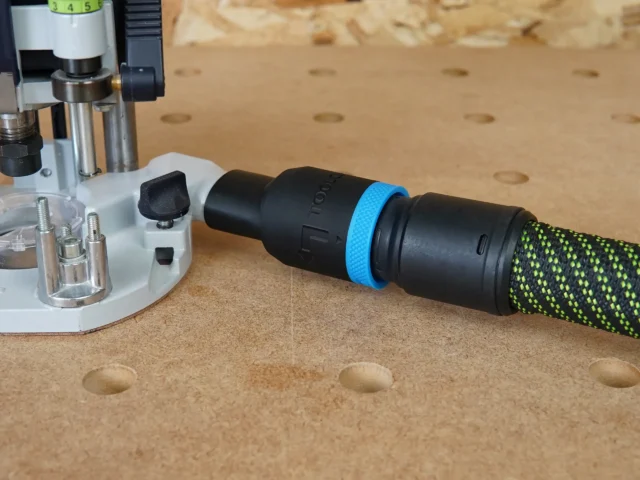 Black hose adapter with blue band attached to Festool 1010 router, connected to green and yellow braided 27mm hose on pegboard workbench