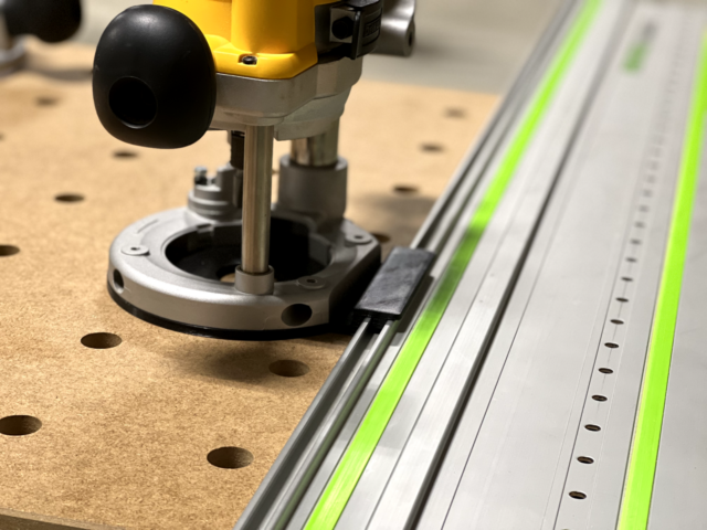 Dewalt DW618 plunge router mounted on a Festool-style guide rail via adapter, showing the black plunge knob and baseplate aligned with the rail's holes
