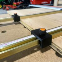 Black plastic parallel guide bracket with orange knurled knob attached to brass Wen track rail on wooden workbench