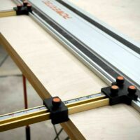 Two black parallel guide brackets with orange knurled knobs mounted on brass Incra T-Track rails attached to a PowerTec track saw rail.