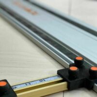 Black parallel guide brackets with orange knobs mounted on a PowerTec track saw rail, positioned on a workbench