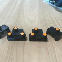 Four black plastic parallel guide brackets with orange knurled knobs, arranged in a row on a wooden surface