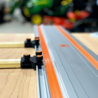 Black plastic parallel guide bracket with orange knurled knobs mounted on Wen track saw rail over wood