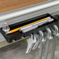 Black storage tray mounted on Festool MFT/3 workbench t-track, holding pencils and pens in tray with four quick clamps hanging from hooks