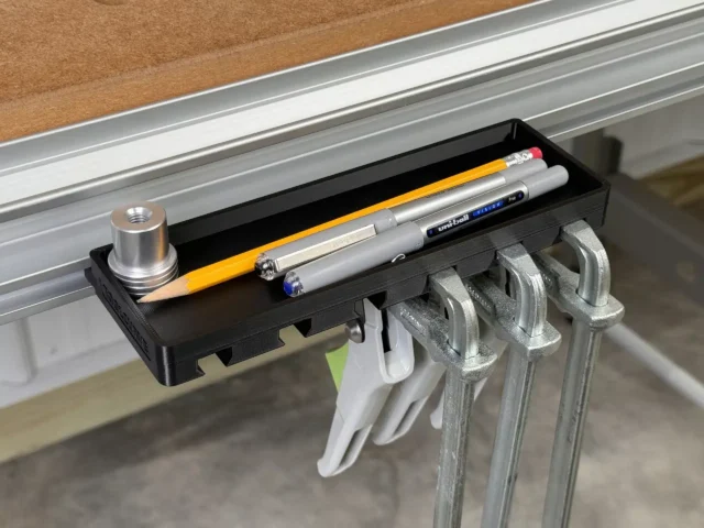 Black storage tray mounted on Festool MFT/3 workbench t-track, holding pencils and pens in tray with four quick clamps hanging from hooks