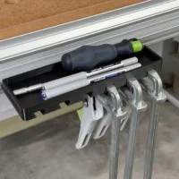 Black clamp rack and storage tray mounted in a Festool MFT table t-track, holding pens, a screwdriver, and clamps