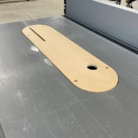 Tan MDF zero clearance insert with slot and mounting holes, positioned on a grey table saw table.