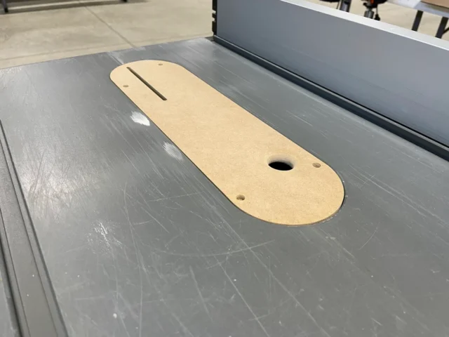 Tan MDF zero clearance insert with slot and mounting holes, positioned on a grey table saw table.
