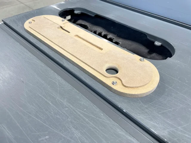 Tan MDF zero clearance insert for Dewalt table saw, shown installed in saw slot with black dust collection shroud visible behind it