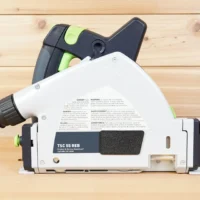 ToolCurve track saw arbor dust cover attached to Festool TS 55, white with black lever and green accents, shown from above on wooden surface