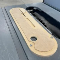 Tan MDF zero clearance insert for Dewalt table saw, displayed in the saw's slot next to black dust collection tube, showing mounting holes and curved edges